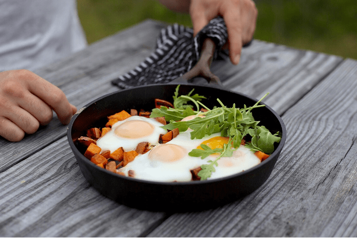 Person preparing a breakfast dish with eggs, sweet potatoes, and greens in a skillet on a wooden table.