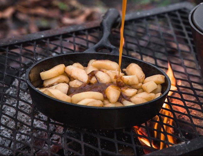 Person pouring a syrup over apples in a cast iron skillet pan over a campfire